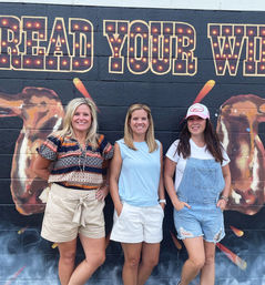 Three smiling women in casual summer outfits pose against a colorful brick-wall mural with marquee-style letters and large painted wings — a playful outdoor street-art photo op