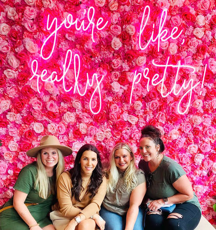 Four smiling women seated in front of a vibrant pink rose wall and neon sign reading "you're like, really pretty!" — playful floral photo wall.