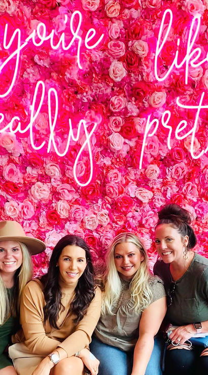Four smiling women seated in front of a vibrant pink rose wall and neon sign reading "you're like, really pretty!" — playful floral photo wall.