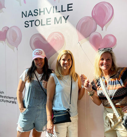 Three smiling women posing with faux balloon strings in front of a pink heart-and-balloon mural that reads “NASHVILLE STOLE MY” — playful Nashville mural photo-op, casual summer outfits and crossbody bags.