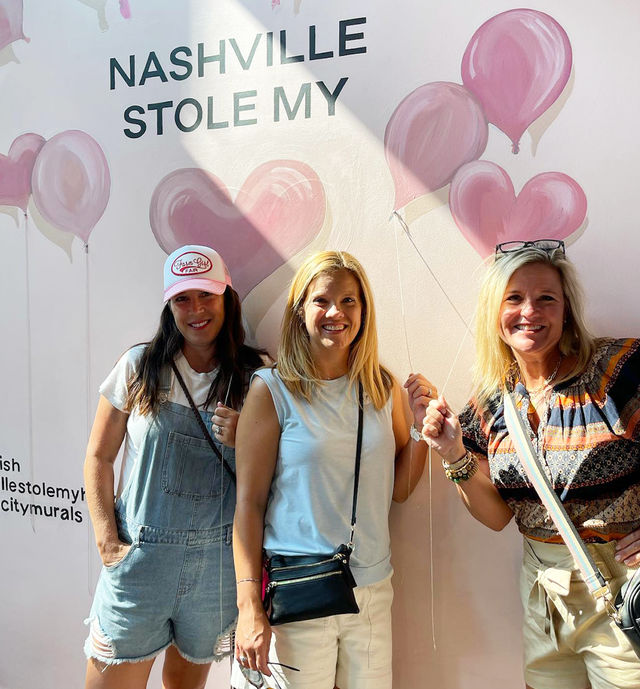 Three smiling women posing with faux balloon strings in front of a pink heart-and-balloon mural that reads “NASHVILLE STOLE MY” — playful Nashville mural photo-op, casual summer outfits and crossbody bags.