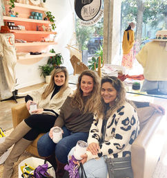 Three smiling women holding insulated tumblers on a couch in a sunlit boutique shop with clothing displays, potted plants, and gift bags