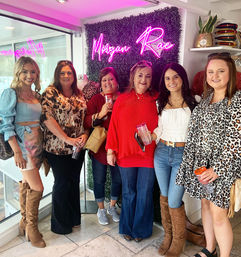 Six smiling women in stylish outfits posing inside a bright women's boutique interior with pink neon lighting, faux greenery wall, large window, and drinks while shopping.
