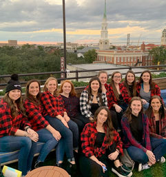 Cheerful group of women in red plaid shirts posing on a Savannah rooftop at sunset with a church steeple and city skyline in the background.