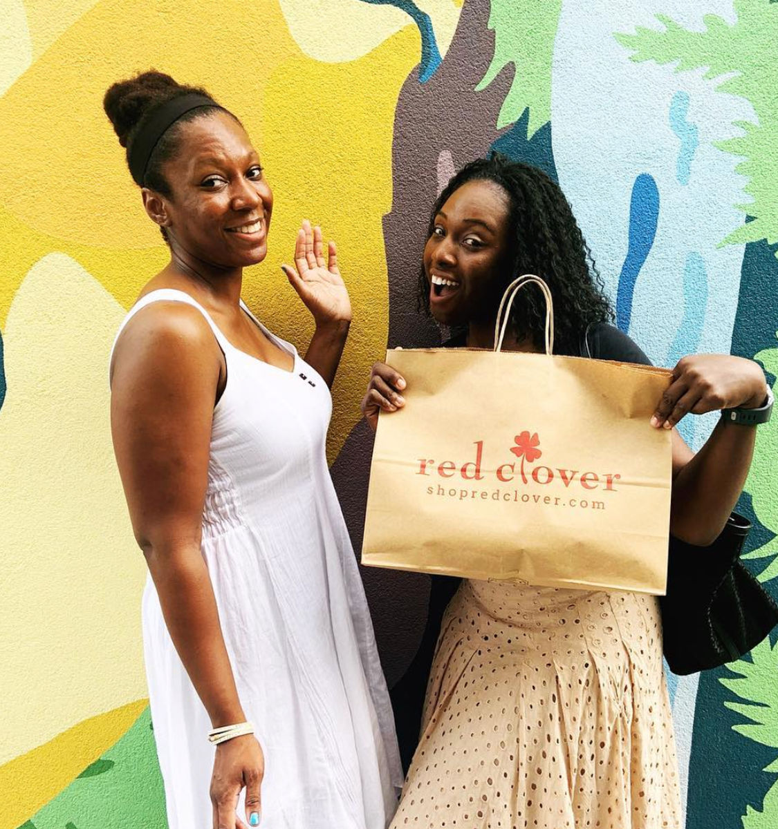 Two smiling women pose in front of a colorful outdoor mural; one waves while the other holds a brown paper shopping bag with a printed logo, capturing a cheerful urban shopping moment.