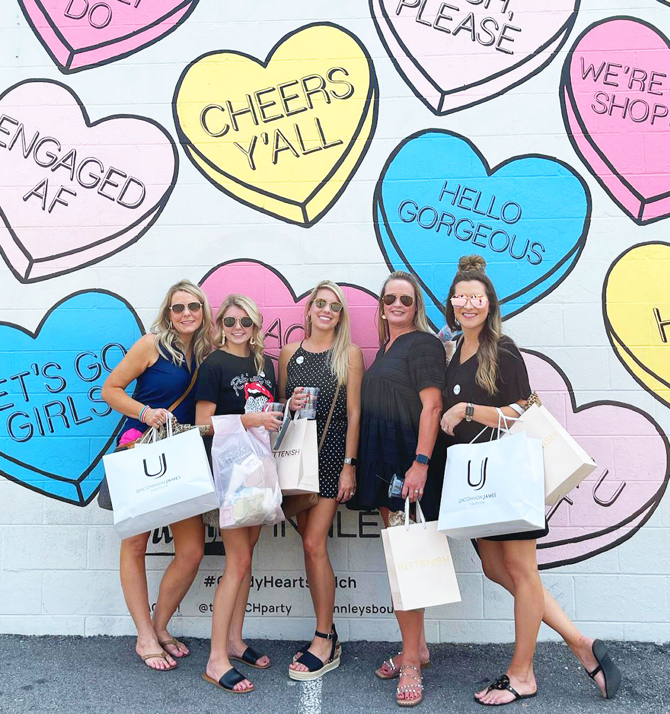 Five friends holding shopping bags pose in front of a pastel conversation-heart mural on a painted brick wall reading phrases like “Cheers y’all” and “Hello Gorgeous.”