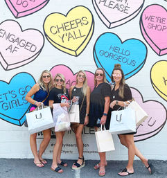 Five friends holding shopping bags pose in front of a pastel conversation-heart mural on a painted brick wall reading phrases like “Cheers y’all” and “Hello Gorgeous.”