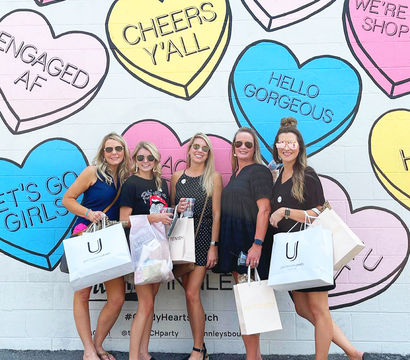 Five friends holding shopping bags pose in front of a pastel conversation-heart mural on a painted brick wall reading phrases like “Cheers y’all” and “Hello Gorgeous.”