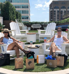 Two women lounging on a sunny urban rooftop patio in white Adirondack chairs, holding drinks with shopping bags on the artificial grass and modern city buildings and blue sky in the background.