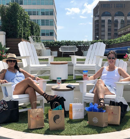 Two women lounging on a sunny urban rooftop patio in white Adirondack chairs, holding drinks with shopping bags on the artificial grass and modern city buildings and blue sky in the background.