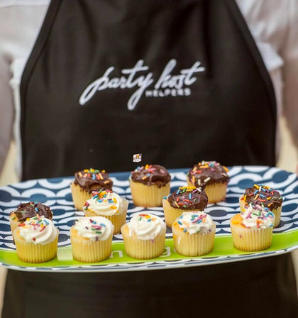 Festive mini cupcakes with chocolate and vanilla frosting and rainbow sprinkles arranged on a blue patterned serving tray held by a person wearing a dark apron — party dessert platter.