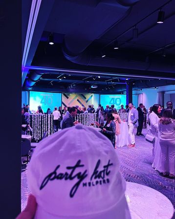 Hand-held white cap with cursive logo in foreground, overlooking a blue-purple lit ballroom of mingling guests, cocktail tables, hanging garlands and large projection screens showing a ‘40 Under 40’ presentation.