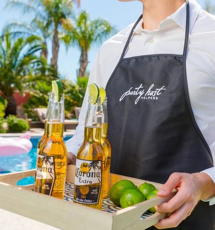 Poolside server in a black apron carrying a wooden tray with three cold beer bottles topped with lime wedges and fresh limes, sunny backyard pool and palm trees in the background.