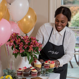 Smiling woman in a black apron arranging frosted cupcakes on a party dessert table with pink and gold balloons and a vase of pink flowers, indoor celebration setup