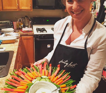 Smiling woman in apron holding a colorful crudités vegetable platter—baby carrots, red pepper strips, green beans, cucumber slices, broccoli and a bowl of creamy dip in a home kitchen; party appetizer, healthy snack.