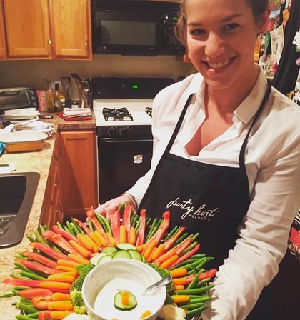 Smiling host in a home kitchen holding a colorful party vegetable platter with baby carrots, green beans, red pepper strips and cucumber rounds arranged around a bowl of creamy dip.