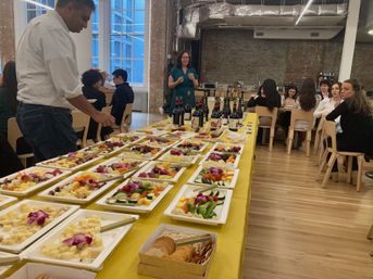 Loft-style office gathering with a long yellow-covered buffet of colorful cheese, fruit, vegetable platters, crackers and wine bottles as guests mingle and sit at tables.