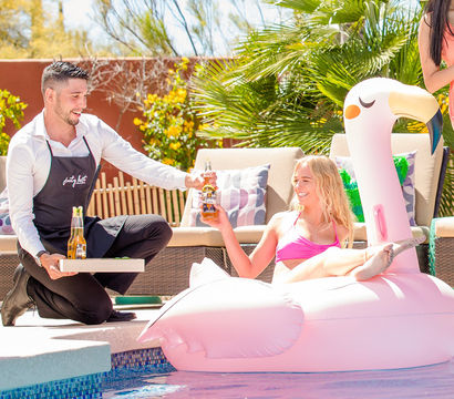 Sunny backyard poolside: woman in a pink bikini lounging on a large pink flamingo float while a server in a black apron kneels at the pool handing her a bottled beer, wicker lounge chairs and palm plants in the background.