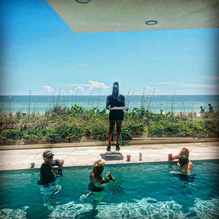 Oceanfront pool scene with three people relaxing with drinks in a pool while a server stands on the deck holding a tray, sandy beach and blue ocean under a sunny sky in the background.