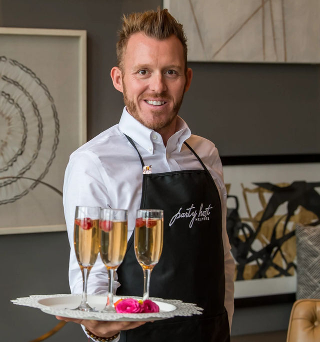 Smiling server in white shirt and black apron holding a tray with three champagne flutes garnished with raspberries and pink roses in a modern indoor party/catering setting.