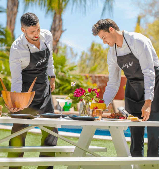Two servers in black aprons arranging plates, wooden bowl, drinks and a vase of pink flowers on a white picnic table at a sunny poolside patio with palm trees