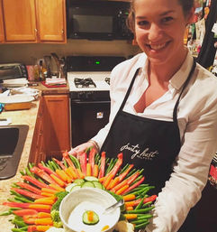 Smiling woman in a home kitchen holding a colorful vegetable party platter — baby carrots, red pepper strips, green beans, cucumber slices and broccoli arranged around a bowl of creamy dip, ready for a casual gathering.