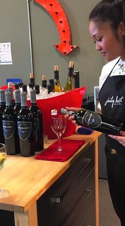 Server in an apron pouring red wine into a glass at an indoor wine-tasting station, row of wine bottles on a wooden counter, ice-filled bucket and sample glasses nearby.