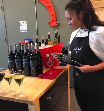 Server in black apron pouring wine at an indoor wine-tasting table—rows of bottled red wine, a red ice bucket with white bottles, and three sample glasses on a wooden counter.