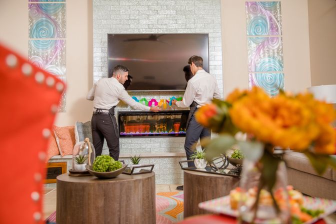 Two people in white shirts and aprons arranging colorful decorations on a modern electric fireplace under a wall-mounted flat-screen TV in a bright contemporary living room with patterned pillows, decorative wall art, and an orange floral centerpiece.