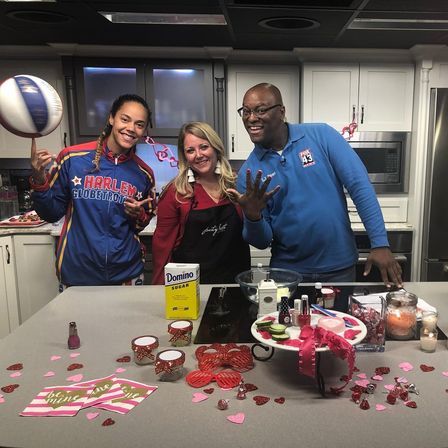 Valentine’s Day studio kitchen segment with three smiling hosts — a Harlem Globetrotter in team jacket spinning a basketball, a woman in an apron, and a man in a blue polo gesturing, countertop scattered with heart confetti, sugar and dessert-making supplies.