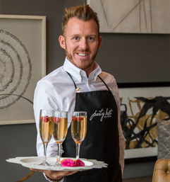 Smiling server in white shirt and black apron holding a tray with three champagne flutes garnished with raspberries and pink flowers, serving at a stylish indoor reception with modern artwork in the background