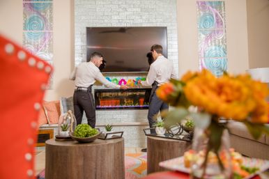 Two people arranging colorful decor on a modern electric fireplace beneath a wall-mounted TV in a bright staged living room with orange flowers in the foreground.