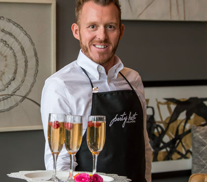 Smiling event server in a white shirt and black apron holding a tray with three champagne flutes garnished with raspberries and a pink rose in a modern indoor reception