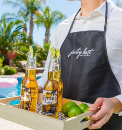 Poolside summer scene: person in a dark apron carries a wooden tray with three chilled beer bottles topped with lime wedges and extra limes, palm trees and pool in the sunny backyard.