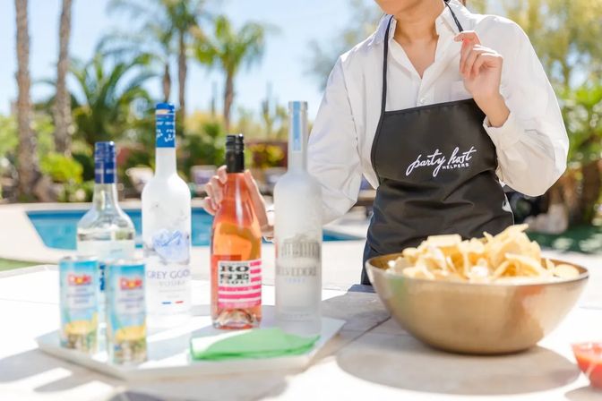 Sunlit poolside party setup with a server in a white shirt and dark apron arranging colorful wine and spirit bottles on a table beside a large bowl of chips, palm trees and a pool in the background.