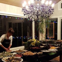 Festive indoor buffet under a sparkling crystal chandelier — chafing dishes, rolls, appetizers and a floral centerpiece as a server prepares food by large windows with colorful string lights.