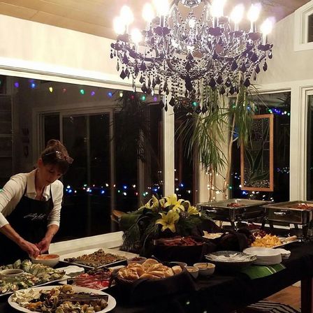 Evening indoor buffet in a cozy dining room — server arranging appetizers on a long table with rolls, platters, chafing dishes, a lily centerpiece, sparkling black chandelier and colorful string lights outside large windows.