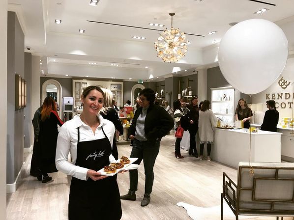 Smiling server in a black apron holding a tray of appetizers inside a bright modern beauty boutique during a store event, shoppers browsing cosmetic displays under a decorative chandelier and oversized white balloon.