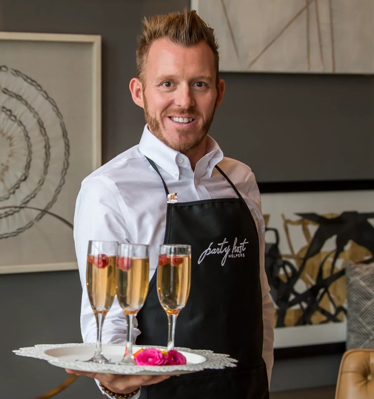 Smiling waiter in a white shirt and black apron holding a tray with three champagne flutes garnished with raspberries and pink flowers, serving sparkling wine at an indoor catered event with modern decor.