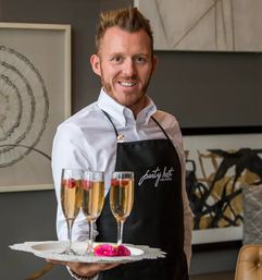 Smiling waiter in a white shirt and black apron holding a tray with three champagne flutes garnished with raspberries and pink flowers, serving sparkling wine at an indoor catered event with modern decor.