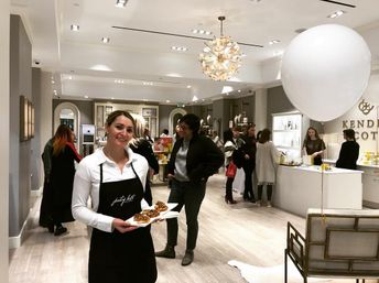 Smiling server holding a tray of appetizers at a chic beauty boutique event, shoppers browse illuminated shelves under a modern chandelier and oversized white balloons.