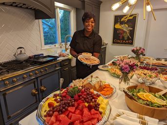 Smiling server in a modern home kitchen holding a tray of crackers and dip next to an island laden with a colorful buffet of fresh fruit, salads, vegetables and appetizers for an indoor dinner party.