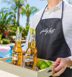 Poolside summer scene: server in a black apron carries a wooden tray with three bottled beers topped with lime wedges and extra limes, palm trees and a swimming pool in the sunny background