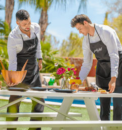 Two men in white shirts and black aprons arranging plates, a wooden salad bowl, cocktails and bright pink flowers on a white picnic table beside a pool in a sunny backyard