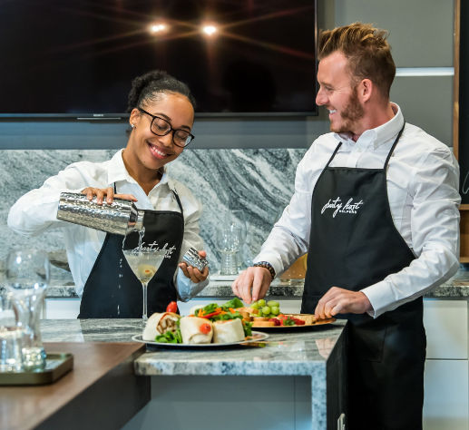 Two smiling staff in black aprons preparing a shaken cocktail and a colorful appetizer platter on a modern marble kitchen island — one pours a drink while the other arranges wraps, grapes, and veggies.