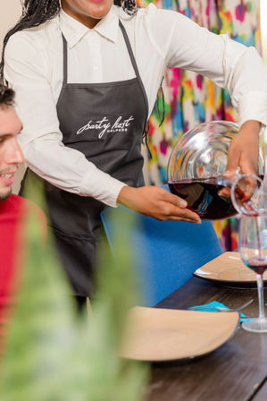 Server in white shirt and dark apron pouring red wine from a glass decanter into a wine glass at a lively, colorfully decorated indoor dinner table — catered dinner service with plates and place settings.