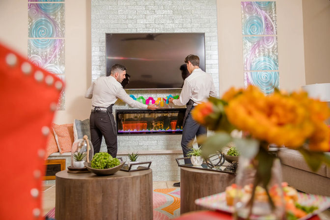 Two staff in white shirts arranging a colorful garland above a modern fireplace and wall-mounted TV in a bright, contemporary living room with orange flowers and succulents in the foreground