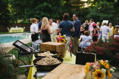 Backyard poolside summer party buffet with skillet of shredded meat and bowls on a yellow table, bright flowers and guests mingling in the garden