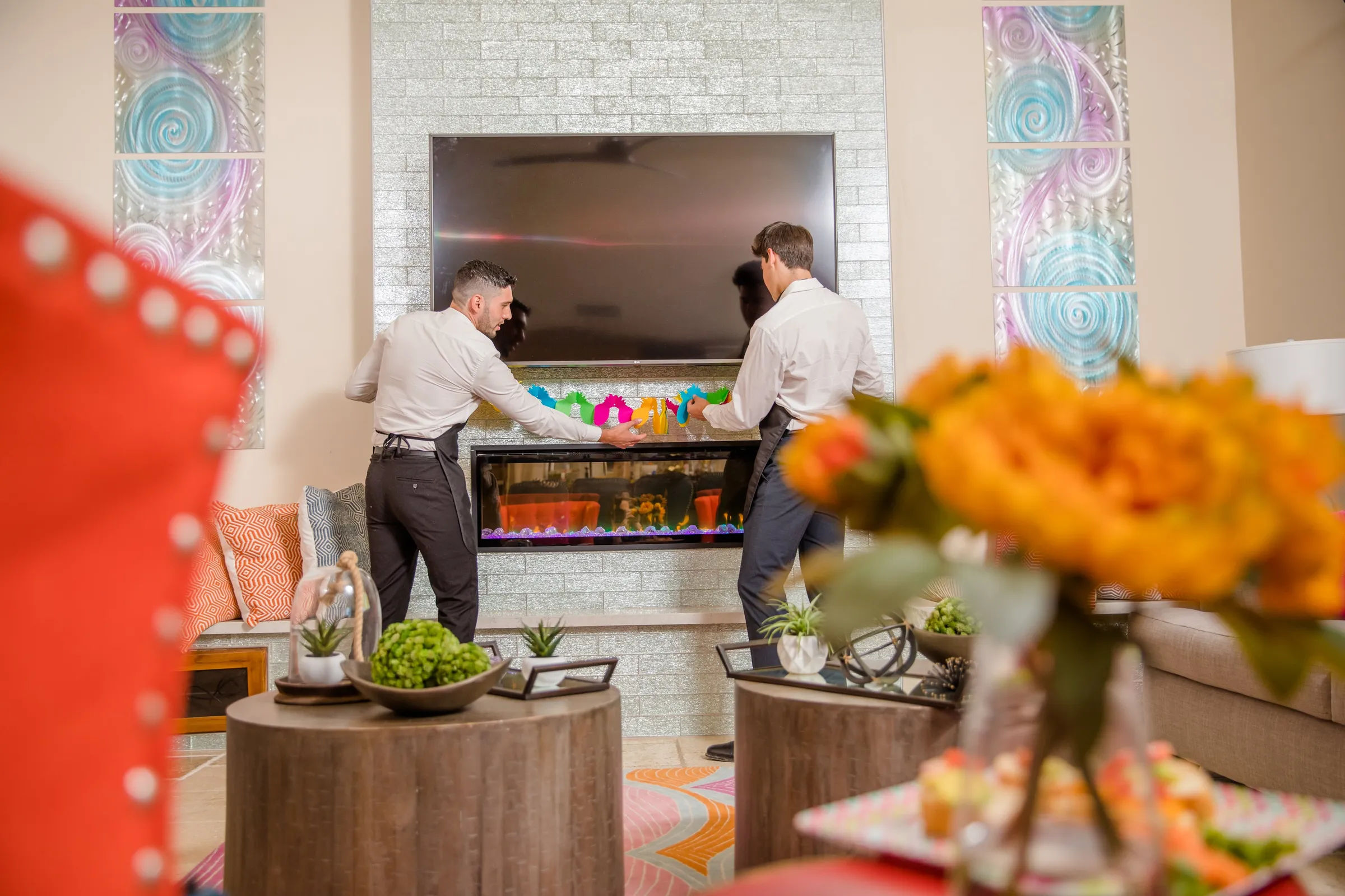 Two people in aprons arranging colorful glassware on a mantel above a modern electric fireplace and wall-mounted TV in a contemporary living room, blurred orange flowers and round wooden coffee tables with succulents in the foreground.