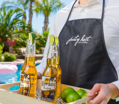 Sunny poolside scene: server in a black apron carrying a wooden tray with three lime-garnished chilled lager bottles and fresh limes, palm trees and backyard pool in the background.
